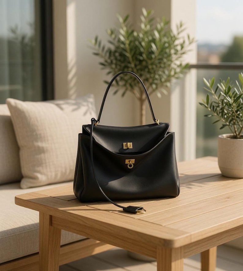 Pu&ograve; includere: Una borsa in pelle nera con manico superiore e finiture color oro. La borsa &egrave; rettangolare con chiusura a patta e una lunga tracolla. &Egrave; appoggiata su un tavolo di legno marrone chiaro, con un cuscino e piante in vaso sullo sfondo.