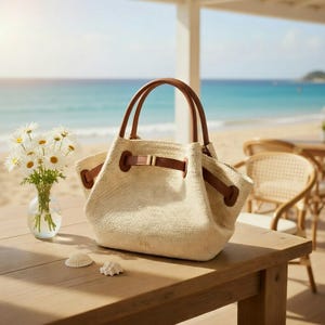 Puede incluir: Un bolso tote tejido beige con asas y detalles de cuero marr&oacute;n. El bolso tiene un cierre de cord&oacute;n y un detalle de hebilla. Un jarr&oacute;n con margaritas y conchas marinas est&aacute; sobre una mesa de madera, con una playa y el mar al fondo.