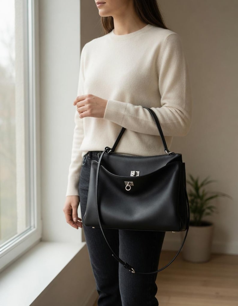 Pu&ograve; includere: Una borsa in pelle nera con manico superiore e chiusura color argento. La borsa ha una forma rettangolare con chiusura a patta e tracolla staccabile. La persona indossa un maglione color crema e jeans scuri.