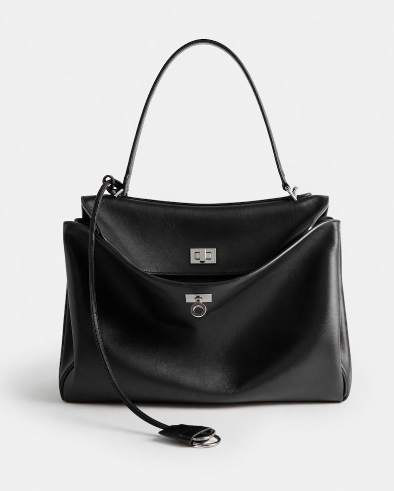 Pu&ograve; includere: Borsa a mano in pelle nera con manico superiore e tracolla staccabile. La borsa presenta una chiusura con patta con chiusura argentata e una tasca frontale con dettaglio ad anello argentato. Design strutturato.