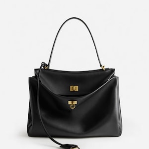 Pu&ograve; includere: Borsa a mano in pelle nera con manico superiore e tracolla staccabile. La borsa presenta dettagli dorati, tra cui una chiusura rettangolare e una chiusura ad anello circolare. Design elegante e minimalista.