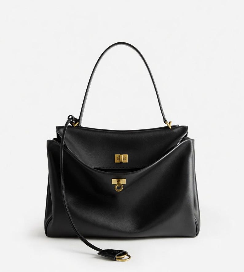 Pu&ograve; includere: Borsa a mano in pelle nera con manico superiore e tracolla staccabile. La borsa presenta dettagli dorati, tra cui una chiusura rettangolare e una chiusura ad anello circolare. Design elegante e minimalista.