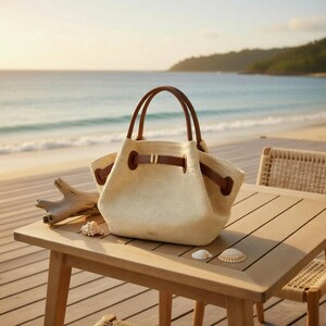 Puede incluir: Un bolso tote tejido beige con asas y detalles de cuero marr&oacute;n est&aacute; sobre una mesa de madera. El bolso tiene un cierre de cord&oacute;n y una peque&ntilde;a hebilla dorada. Conchas marinas y madera flotante tambi&eacute;n est&aacute;n sobre la mesa, con una playa y el oc&eacute;ano al fondo.
