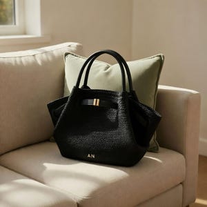 Puede incluir: Bolso tote negro tejido con asas negras y cierre de hebilla dorada. El bolso tiene las iniciales "AN" en letras doradas. Est&aacute; sobre un sof&aacute; beige con un coj&iacute;n verde.