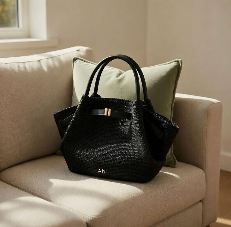 Puede incluir: Bolso tote negro tejido con asas negras y cierre de hebilla dorada. El bolso tiene las iniciales "AN" en letras doradas. Est&aacute; sobre un sof&aacute; beige con un coj&iacute;n verde.