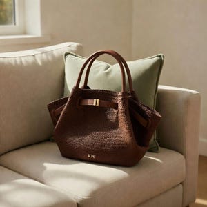 Puede incluir: Un bolso tote tejido marr&oacute;n con asas de cuero y cierre de correa de cuero. El bolso tiene una hebilla dorada y las iniciales "AN" son visibles en la parte delantera. El bolso est&aacute; sobre un sof&aacute; beige con un coj&iacute;n verde.