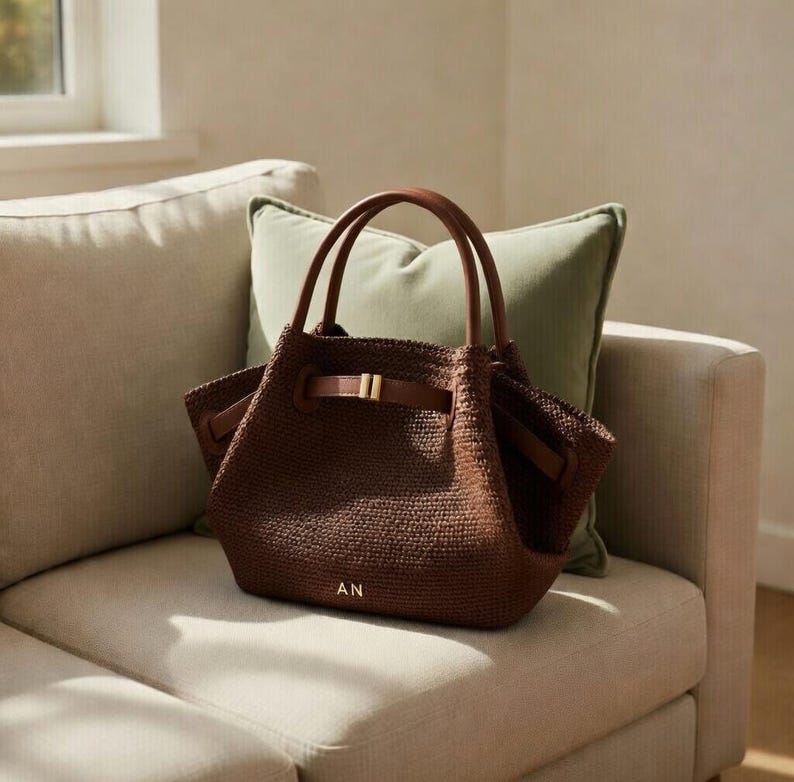 Puede incluir: Un bolso tote tejido marr&oacute;n con asas de cuero y cierre de correa de cuero. El bolso tiene una hebilla dorada y las iniciales "AN" son visibles en la parte delantera. El bolso est&aacute; sobre un sof&aacute; beige con un coj&iacute;n verde.
