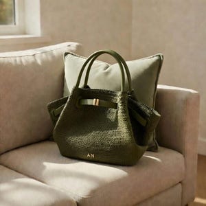 Puede incluir: Bolso tote tejido verde oliva con asas a juego y hebilla dorada. El bolso presenta las iniciales "AN" en letras doradas. El bolso est&aacute; sobre un sof&aacute; beige con un coj&iacute;n verde.
