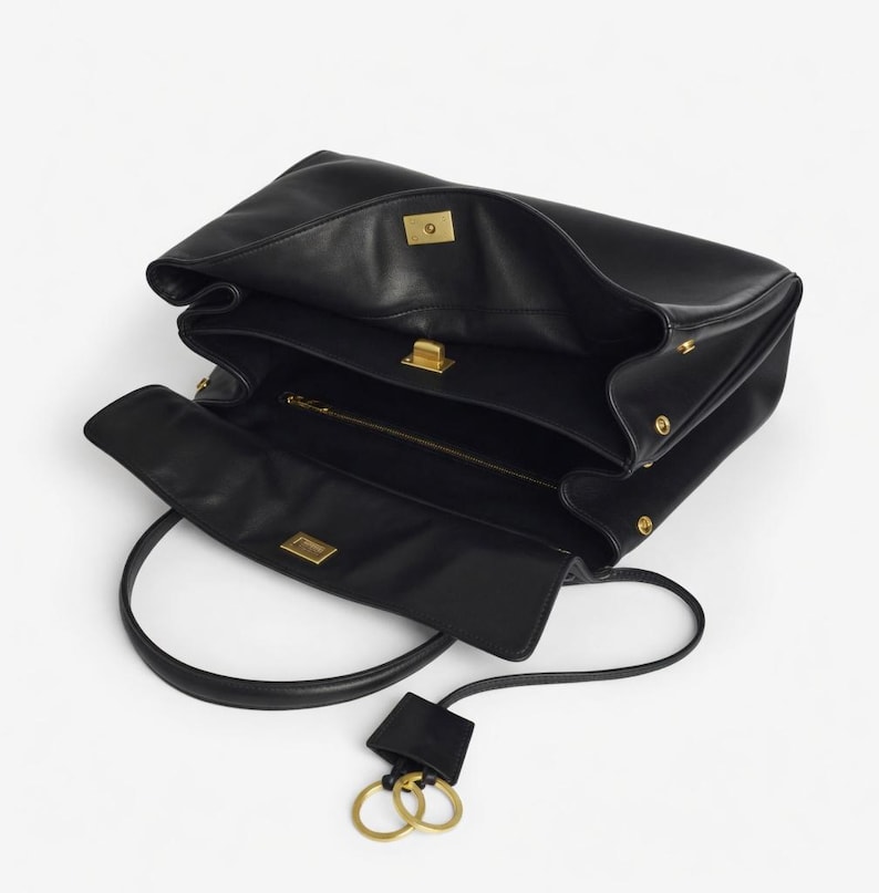 Pu&ograve; includere: Borsa in pelle nera dal design strutturato. La borsa presenta un manico superiore, una tracolla staccabile e finiture dorate. L'interno &egrave; visibile, rivelando pi&ugrave; scomparti e una tasca con cerniera. Un portachiavi con due anelli dorati pende dalla borsa.
