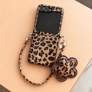 May include: A phone case featuring a leopard print design, complete with a matching strap and a flower-shaped charm. The case is open, displaying the phone screen. The strap and charm also have the leopard pattern. The case is designed for phone protection.