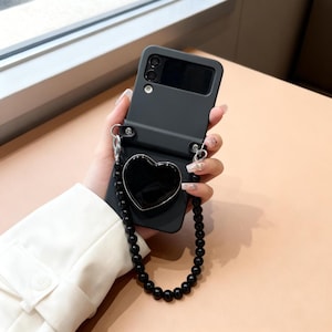 Op de afbeelding: Zwarte telefoonhoes voor een klaptelefoon, met een hartvormige grip en een kralenriem. De hoes heeft uitsparingen voor de camera en het scherm. De riem is gemaakt van zwarte kralen en zilverkleurige hardware.