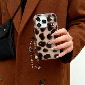 May include: Clear phone case with a brown and tan animal print design. The case has a beaded wrist strap with brown, tan, and clear beads. The case has a camera cutout and is held against a brown coat.