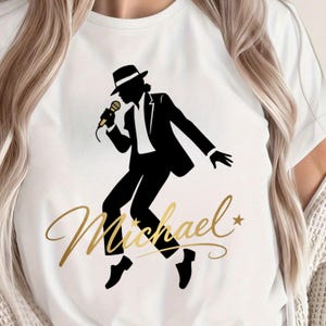 May include: White t-shirt featuring a black silhouette of a singer in a suit and hat, holding a microphone. The name "Michael" is written in gold script below the figure, with a star accent.