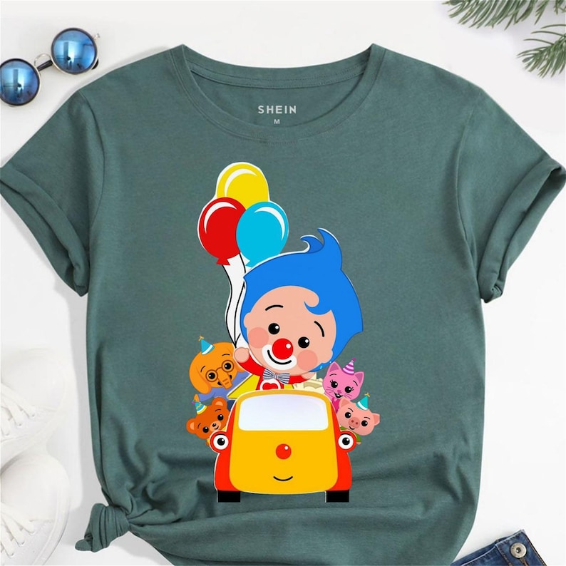 Op de afbeelding: Groen t-shirt met een cartoon clown afbeelding met ballonnen. Het ontwerp toont een gele auto en cartoon dieren. De merknaam "SHEIN" staat op het t-shirt. Een casual kledingstuk.