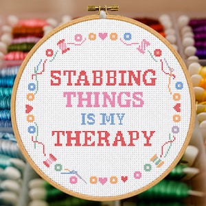 May include: A circular wooden embroidery hoop containing white fabric with a cross-stitch design. The text reads STABBING THINGS IS MY THERAPY in red, pink, and blue. A border of small colorful sewing icons surrounds the text.