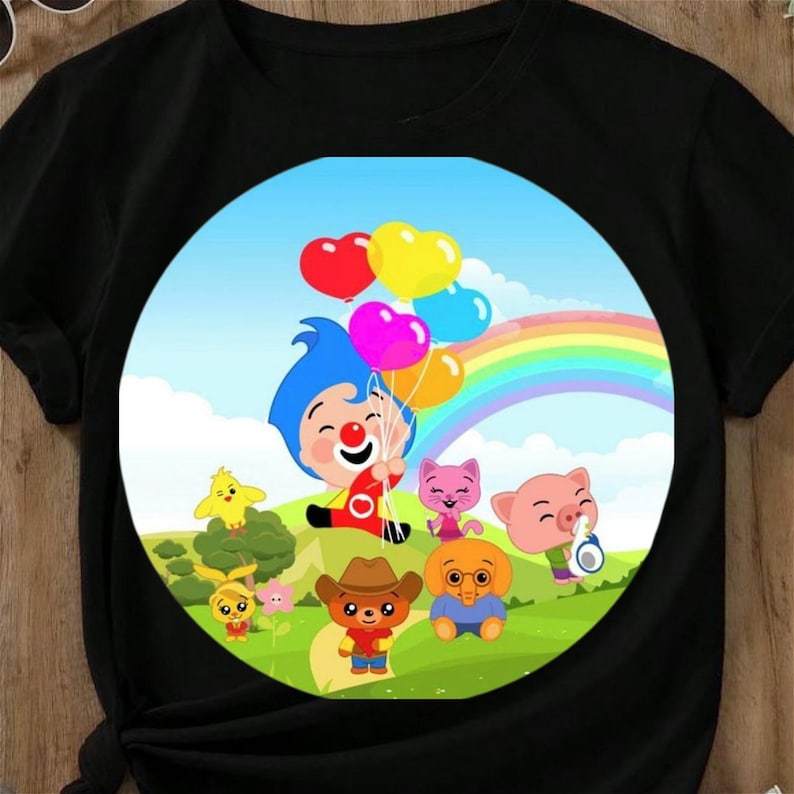 Op de afbeelding: Zwart T-shirt met een kleurrijke cartoon illustratie. Het ontwerp bevat een clown met hartvormige ballonnen, een regenboog en diverse cartoon dieren. De achtergrond is een blauwe lucht en groen gras.