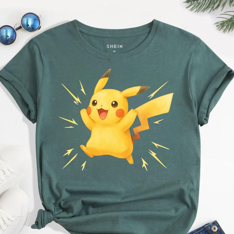 K&ouml;nnte beinhalten: Petrolfarbenes T-Shirt mit einem gelben Pikachu-Motiv und gelben Blitzen. Das Shirt hat hochgekrempelte &Auml;rmel und den Markennamen "SHEIN" oben in der Mitte aufgedruckt. Sonnenbrille und wei&szlig;e Schuhe sind im Hintergrund zu sehen.