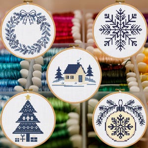 May include: Five circular embroidery hoops featuring cross-stitch designs in blue and cream. The patterns include a wreath with a bow, a large snowflake, a house with pine trees, a decorated Christmas tree with gifts, and a snowflake ornament.