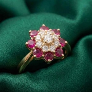 May include: A gold ring featuring a floral design with a cluster of small, round, red gemstones surrounding a center of white diamonds. The ring is set against a dark green, silky background.