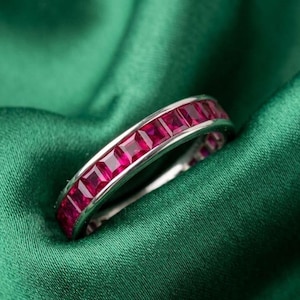 Puede incluir: Un anillo de plata con una fila continua de piedras preciosas rojas vibrantes, de corte cuadrado. El anillo se presenta sobre un fondo de tela verde oscuro, creando un contraste llamativo. Las piedras preciosas están espaciadas uniformemente.