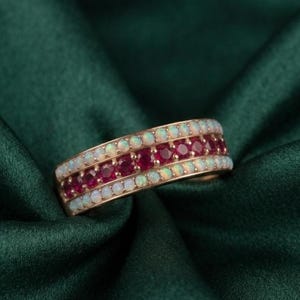 May include: A gold band ring featuring three parallel rows of gemstones. The outer rows consist of small, round, iridescent white opals, while the center row is composed of vibrant, round, deep red rubies set in a polished metal band.