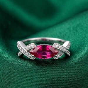 Op de afbeelding: Een zilverkleurige ring met een centrale, in marquise geslepen, levendige roze-rode edelsteen. De ringband is ontworpen met een overlappend kruispatroon aan beide zijden, ingelegd met kleine, heldere, fonkelende pavé-gezette steentjes.