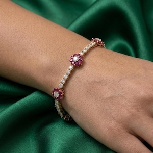 Puede incluir: Una pulsera dorada con piedras claras y de color rubí alternadas. La pulsera presenta tres racimos de rubíes en forma de flor. La pulsera se lleva en la muñeca sobre un fondo de satén verde.