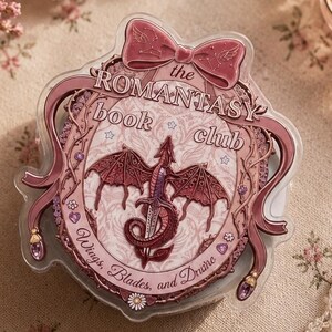 May include: A clear, decorative badge with a pink bow and the text "The Romantasy Book Club." The badge features a dragon design with the words "Wings, Blades, and Desire."