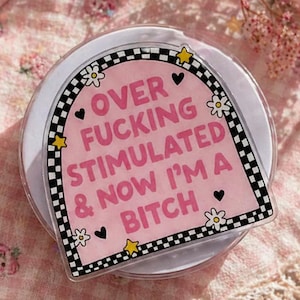 May include: A pink and white phone grip with the text "OVER FUCKING STIMULATED & NOW I'M A BITCH" in a black and white checkered border. The grip has a white base and is decorated with small hearts, stars, and flowers.