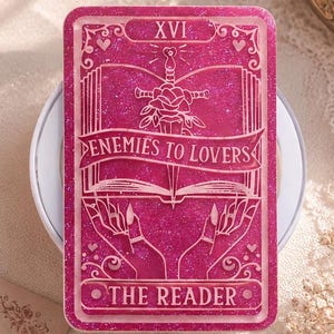 May include: A magenta-colored tarot card with glitter, featuring the text "XVI" at the top. The card design includes a sword through a rose, an open book, and the words "ENEMIES TO LOVERS" and "THE READER".