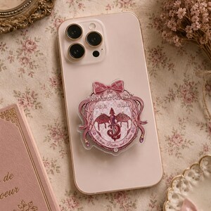 May include: A pink smartphone with a decorative pop-socket featuring a dragon design and the text "The Romantasy Book Club". The pop-socket has a bow at the top and is surrounded by a decorative border. The phone is on a floral patterned surface.