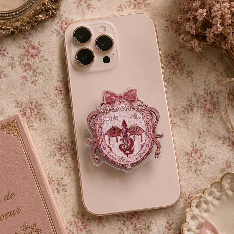 May include: A pink smartphone with a decorative pop-socket featuring a dragon design and the text "The Romantasy Book Club". The pop-socket has a bow at the top and is surrounded by a decorative border. The phone is on a floral patterned surface.