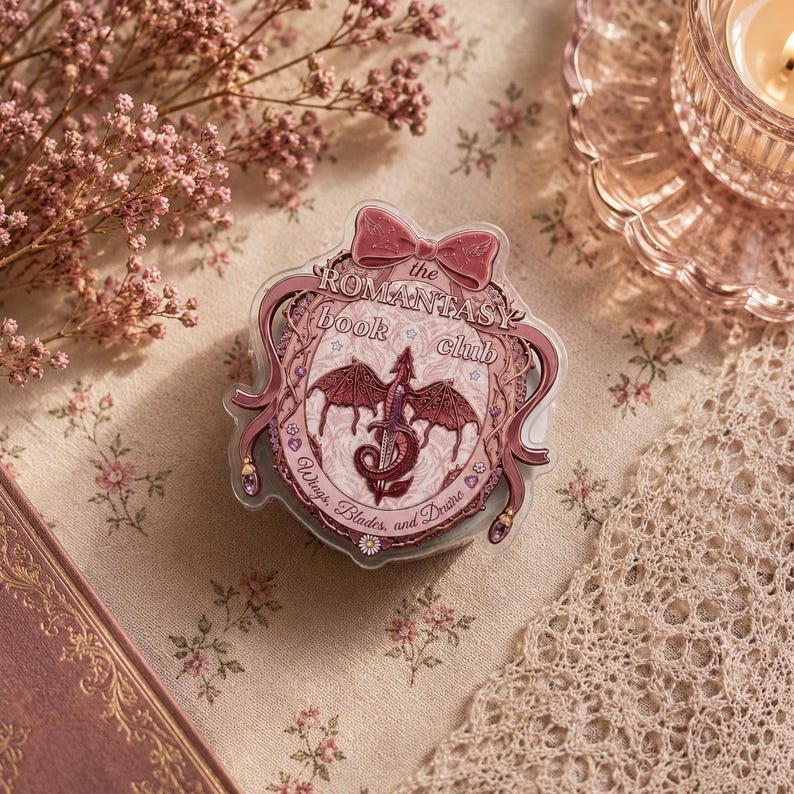 May include: A clear, decorative badge with a pink bow and the text "The Romantasy Book Club." The badge features a dragon design with the words "Wings, Blades, and Desire."