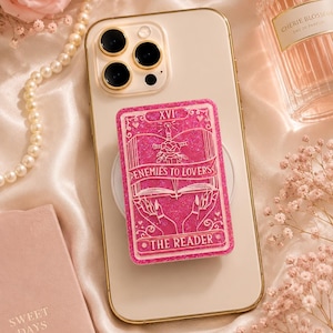 May include: A pink glittery phone grip featuring a tarot card design. The card reads "Enemies to Lovers" and "The Reader." The grip is attached to a gold-rimmed phone. Other items in the image include a pearl necklace and a perfume bottle.
