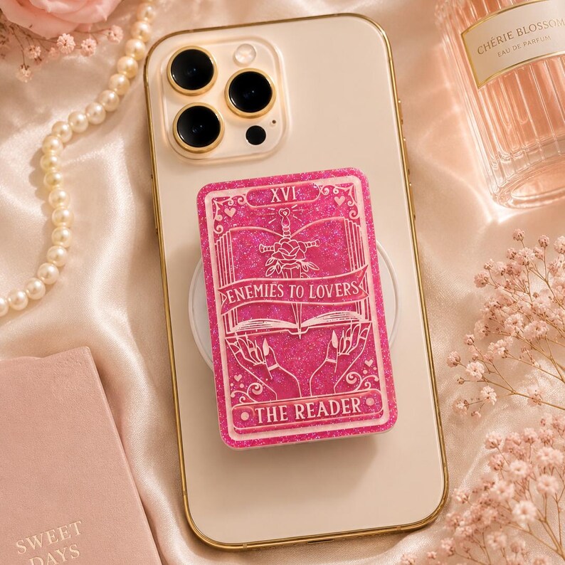 May include: A pink glittery phone grip featuring a tarot card design. The card reads "Enemies to Lovers" and "The Reader." The grip is attached to a gold-rimmed phone. Other items in the image include a pearl necklace and a perfume bottle.