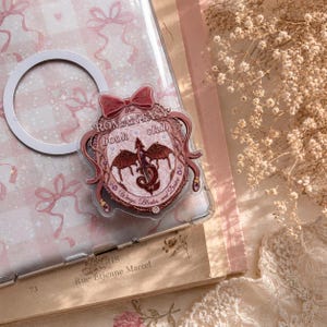 May include: A clear phone case with a decorative emblem featuring a dragon, bow, and text that reads "The Romantas Book Club". The case is on a pink and white patterned surface with a white ring.