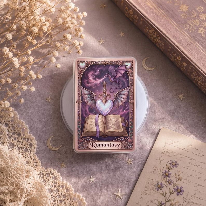 May include: A rectangular card with a fantasy illustration featuring a heart-shaped crystal, a sword, and a dragon. The card has the word "Romantasy" at the bottom. The color palette includes shades of purple, pink, and white.