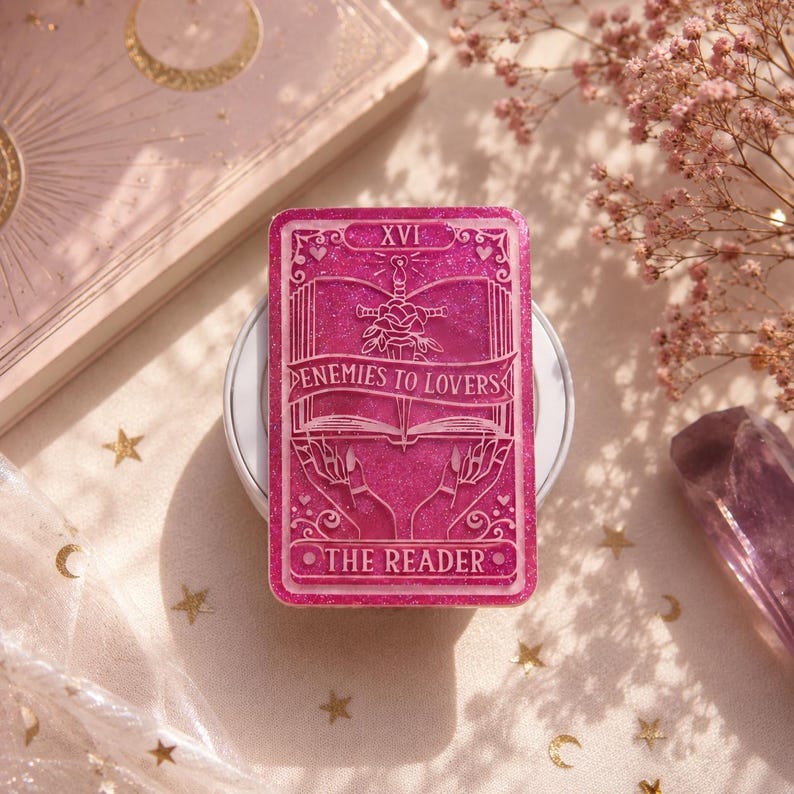 May include: A pink, glittery tarot card with the text "ENEMIES TO LOVERS" and "THE READER". The card features a sword, a book, and hands. The card is set on a white surface, with a tarot deck and crystals in the background.