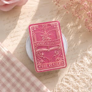 May include: A pink tarot card with the text "ENEMIES TO LOVERS" and "THE READER". The card features a sword, rose, and open book design. The card is set on a white wireless charger.