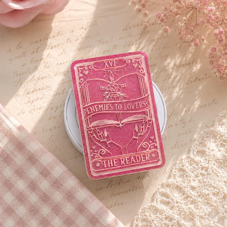 May include: A pink tarot card with the text "ENEMIES TO LOVERS" and "THE READER". The card features a sword, rose, and open book design. The card is set on a white wireless charger.