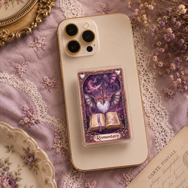 May include: A phone with a pop-up grip featuring a fantasy illustration. The artwork includes a heart pierced by a sword, wings, a dragon, and an open book. The word "Romantasy" is printed at the bottom.