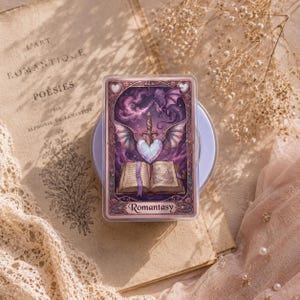 May include: A rectangular card with a fantasy illustration featuring a dragon, sword, and heart. The card has a purple and pink color scheme with the word "Romantasy" at the bottom. The card is set on a light purple base, with a vintage book and lace in the background.