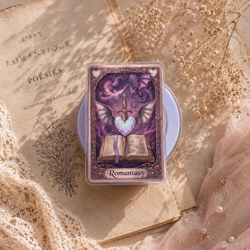 May include: A rectangular card with a fantasy illustration featuring a dragon, sword, and heart. The card has a purple and pink color scheme with the word "Romantasy" at the bottom. The card is set on a light purple base, with a vintage book and lace in the background.
