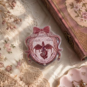 May include: A clear acrylic pin with a pink bow and the words "the ROMANTASY book club". The pin features a red dragon and the words "Wings, Blades, and Drune". The pin is on a lace surface with a book and a pink dish.