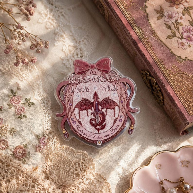 May include: A clear acrylic pin with a pink bow and the words "the ROMANTASY book club". The pin features a red dragon and the words "Wings, Blades, and Drune". The pin is on a lace surface with a book and a pink dish.