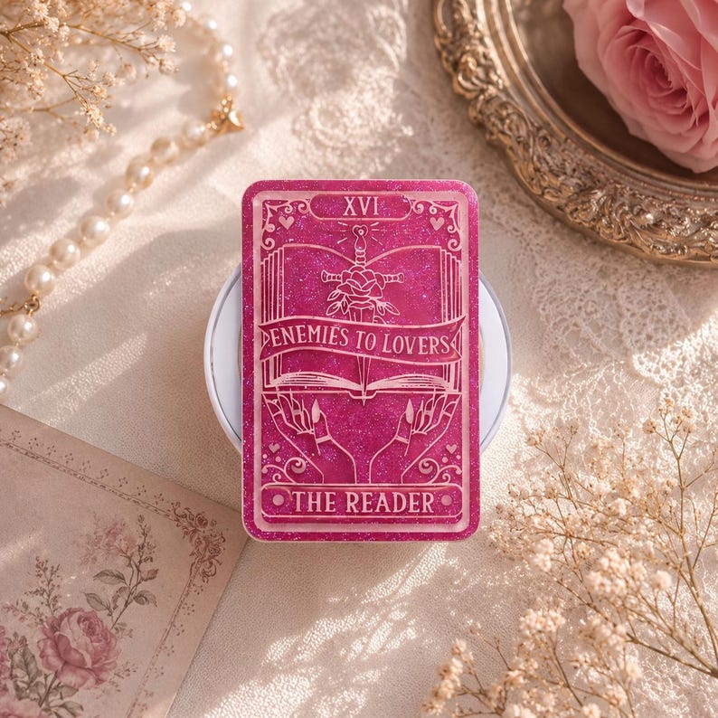 May include: A magenta-colored tarot card with glitter, featuring the text "XVI" at the top. The card design includes a sword through a rose, an open book, and the words "ENEMIES TO LOVERS" and "THE READER".