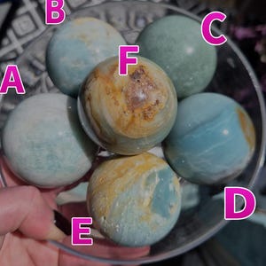 May include: A collection of five smooth, polished stone spheres in a clear bowl, labeled with pink letters A through F. The spheres feature marbled patterns in shades of light blue, teal, cream, and brown, with varying levels of color saturation.