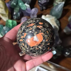 Peut inclure: Une pierre sphérique polie présentant une couleur de base noir profond avec des inclusions tachetées orange et vert clair. La surface affiche un réseau complexe de fines veines blanches et de taches irrégulières, créant un motif contrasté.