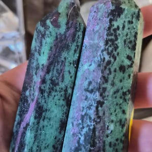 May include: Two polished crystal towers featuring a mottled pattern of vibrant green, deep black, and magenta-pink mineral inclusions. Each stone is shaped into a hexagonal prism with a pointed top, displaying a smooth, reflective surface texture.