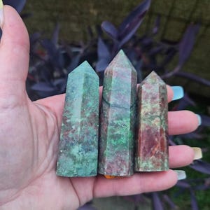 May include: Three polished crystal towers with hexagonal points, featuring a mottled blend of forest green, deep red, and earthy brown mineral inclusions. The stones display a smooth, reflective surface with natural variations in color and texture.